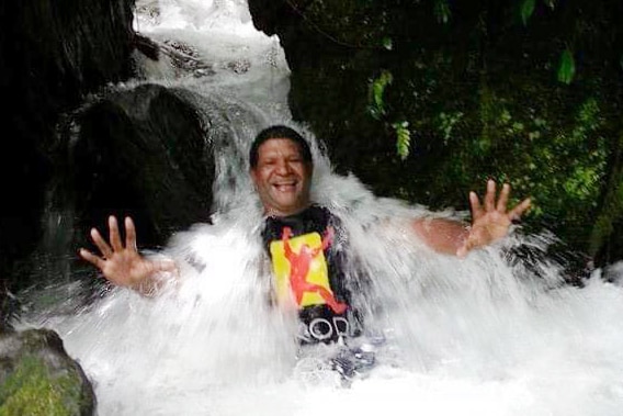 You see a man in a waterfall smiling being inundated by water with his palms outstretched to the camera.