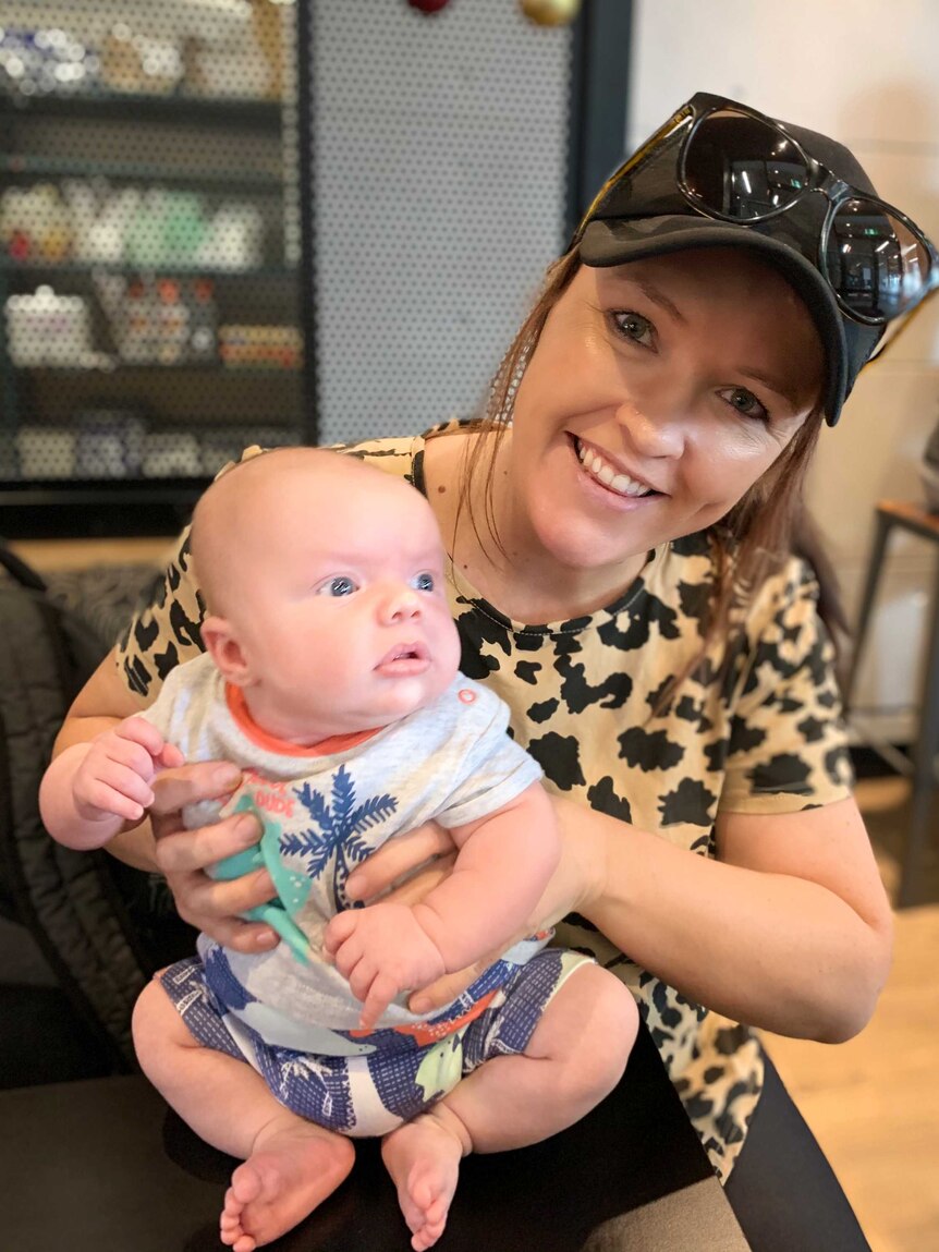 Baby Hamish is held by his mum Julia McLennan. Julia is smiling at the camera and wearing a black hat and sunglasses.