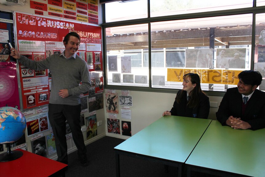 Man points at a poster and two school kids sit at desks