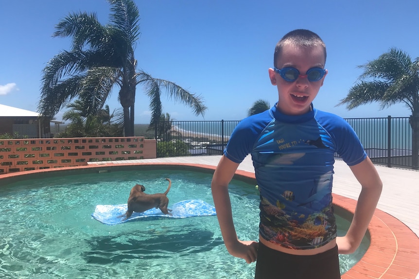 A boy wearing goggles, blue swimming shirt, pool, dog, palm trees, ocean in background.
