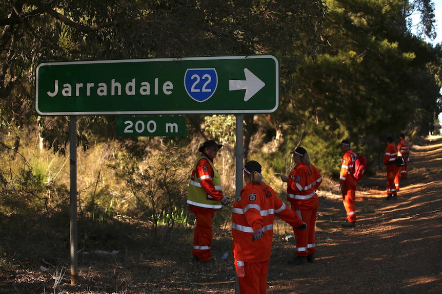 A row of SES volunteers in hi-vis clothing line the side of Albany Highway searching near a sign pointing towards Jarrahdale.
