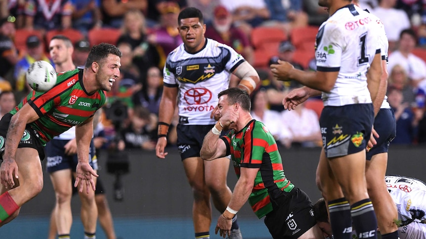 An NRL player flings the ball away in triumph after scoring a try as his teammate looks on.