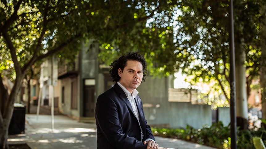 The artist, wearing a navy blue suit an white shirt, sits on a park bench in a leafy inner-Sydney suburb.