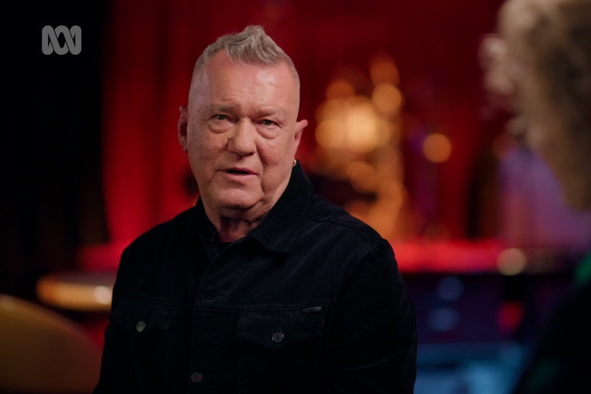 Jimmy Barnes mid sentence on the left of the picture wearing a black top with short grey hair, the background is out of focus