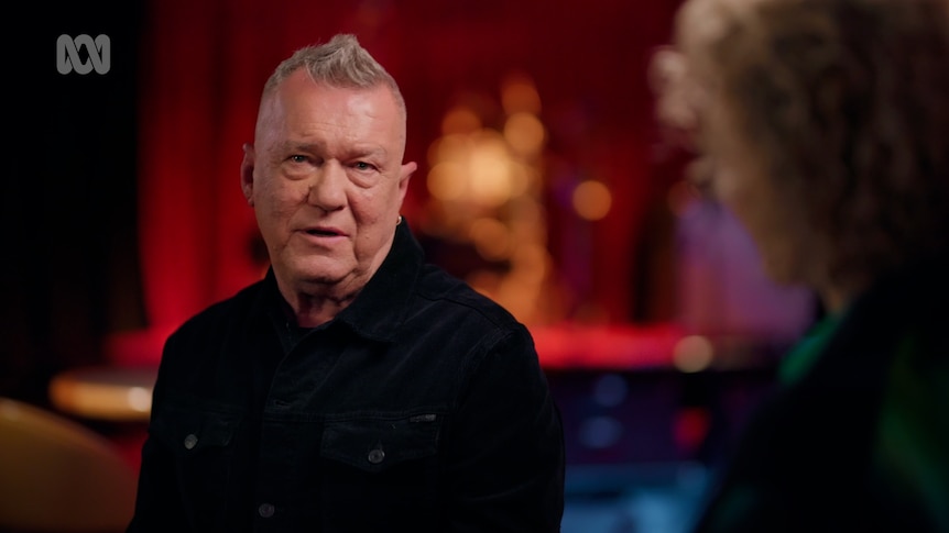 Jimmy Barnes mid sentence on the left of the picture wearing a black top with short grey hair, the background is out of focus