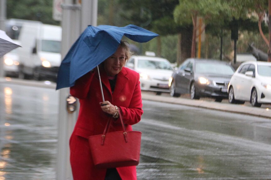 A woman wearing a red coat walks along a footpath carrying a buckling umbrella above her head with traffic in the background.