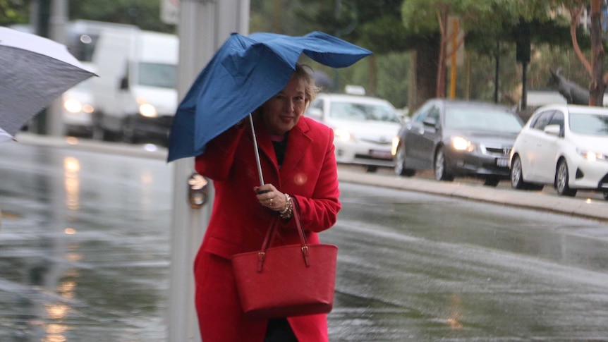 A woman wearing a red coat walks along a footpath carrying a buckling umbrella above her head with traffic in the background.