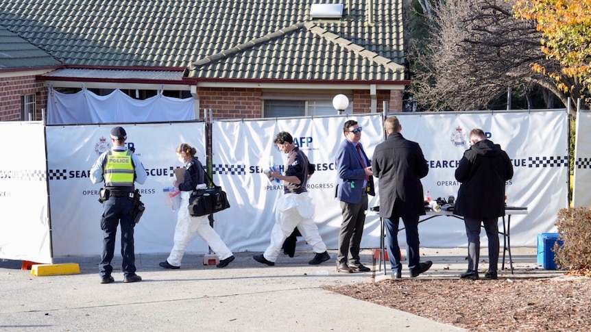 Police officers and forensic officers walking into a house surrounded with blue police tape.