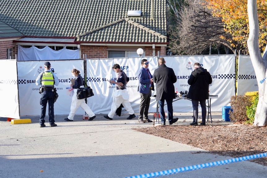 Police officers and forensic officers walking into a house surrounded with blue police tape.
