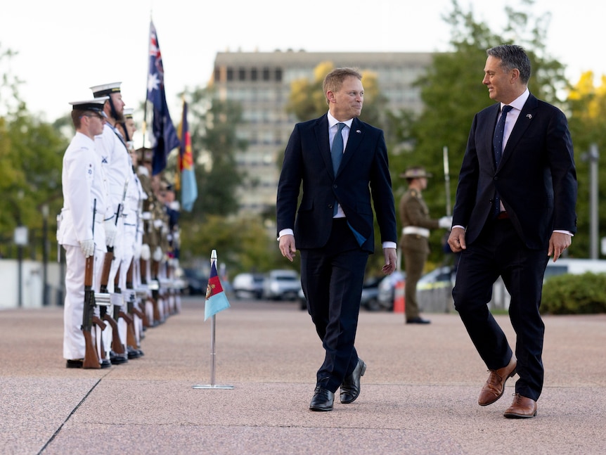 Two men in suits walk together past soldiers standing to attention in a ceremony