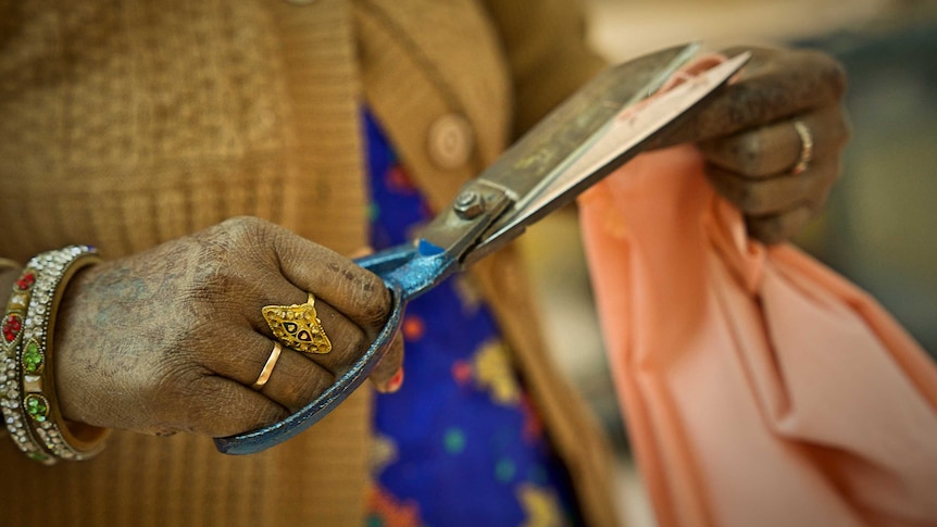 A woman is seen wearing a yellow cardigan with bracelets on her wrist and rings on her fingers as she cuts fabric.