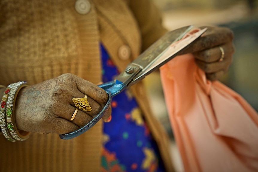 A woman is seen wearing a yellow cardigan with bracelets on her wrist and rings on her fingers as she cuts fabric.