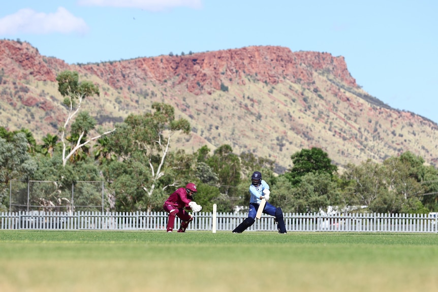 Two cricket players in action on an oval, on a sunny day, with red rocky mountains in the background.