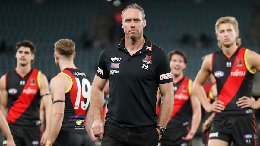 Essendon coach Ben Rutten looks stony-faced as his dejected players stand behind him after a big loss.