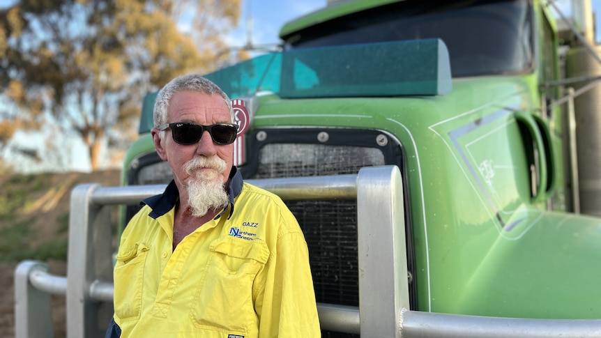 A man with yellow shirt, grey hair and beard stands in front of his truck wearing sunglasses.