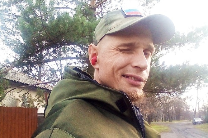 A man in a military uniform smiles winks at the camera. A Russian flag is visible on his hat.