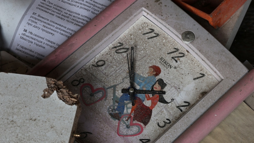 A clock with art of a couple riding a bike lays among the rubble. 