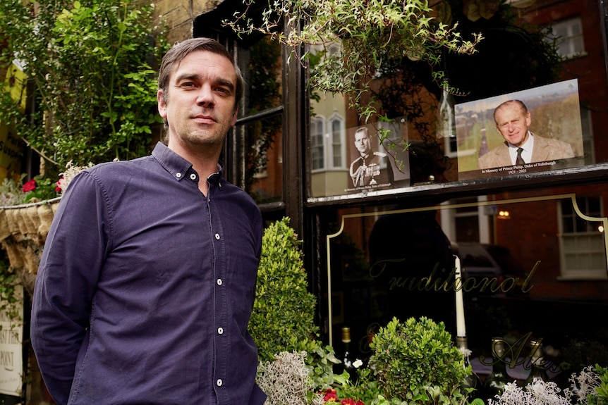 A man in a blue shirt stands next to a pub window containing Prince Philip portraits.