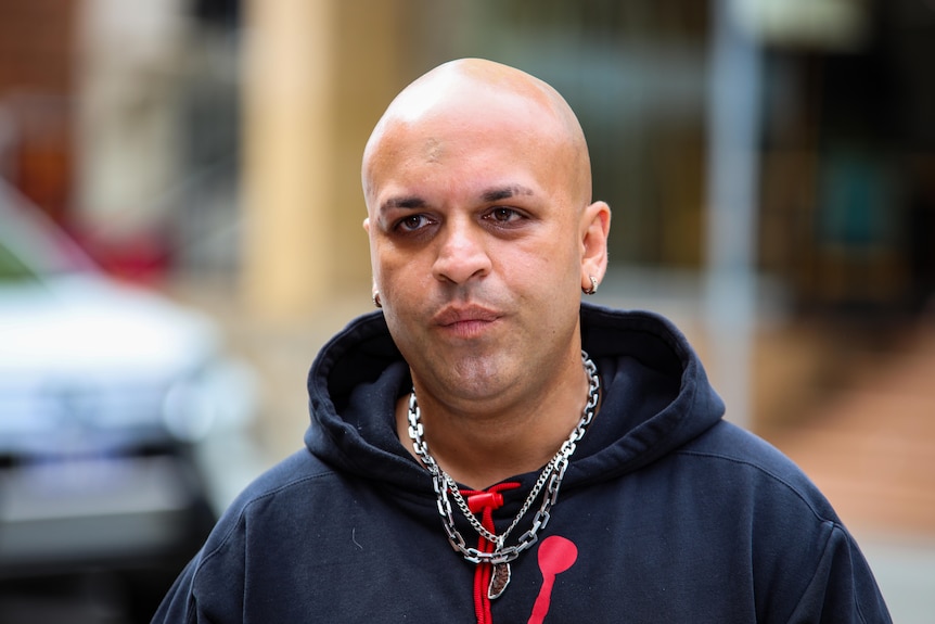 A close-up photo of a man wearing a blue jumper and several necklaces.