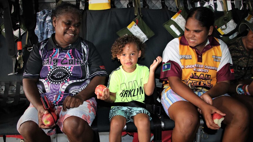 An Aboriginal woman, child and teenager each holding apples and smiling in a military plane