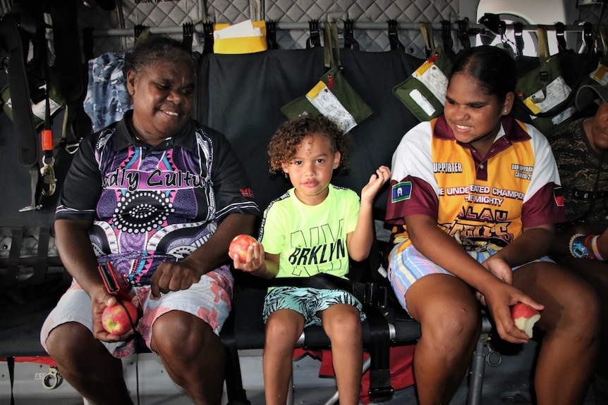 An Aboriginal woman, child and teenager each holding apples and smiling in a military plane