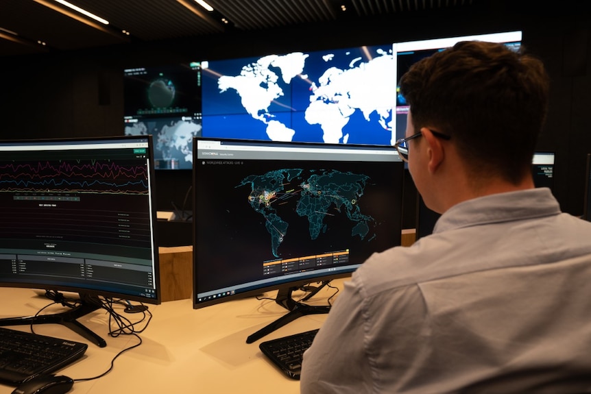A man in a business shirt sits at a desk in a room full of computers looking at a world map on the screen.