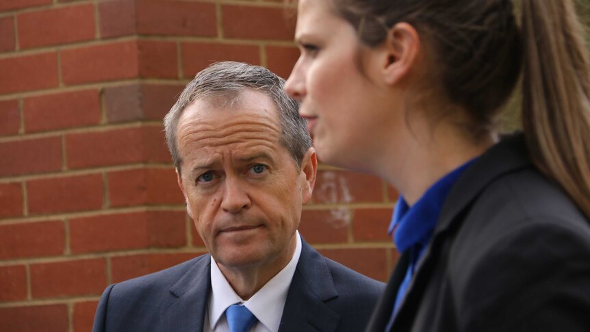 Bill Shorten looks at a woman speaking, in front of a brick wall.