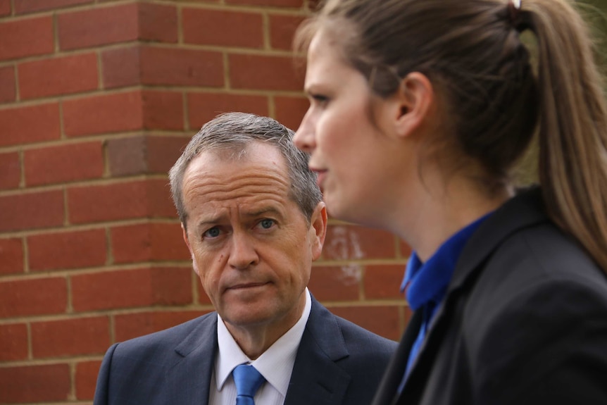 Bill Shorten looks at a woman speaking, in front of a brick wall.