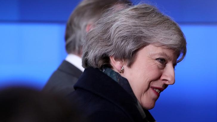 Theresa May looks to her right in image taken from the side. She wears a dark coat against a blue background.