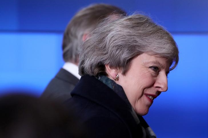 Theresa May looks to her right in image taken from the side. She wears a dark coat against a blue background.