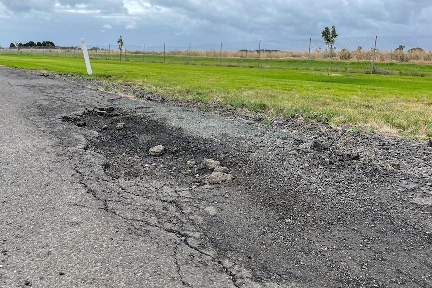 Cracked and potholed road with grass and field visible behind a wire fence