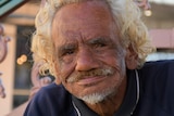 An Aboriginal man with curly blonde hair smiles at the camera.