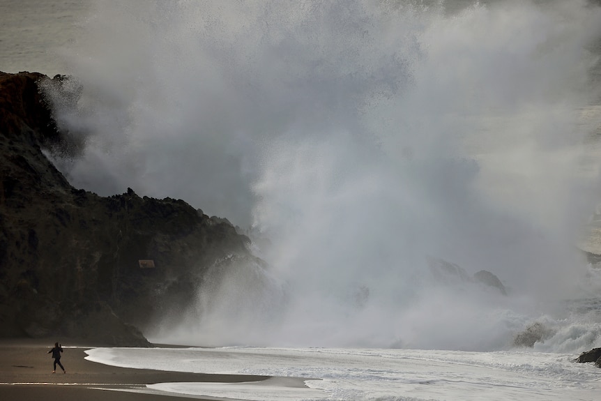 A person watches a huge wave smash into a coastal rock, turning into white mist that rises into the sky.