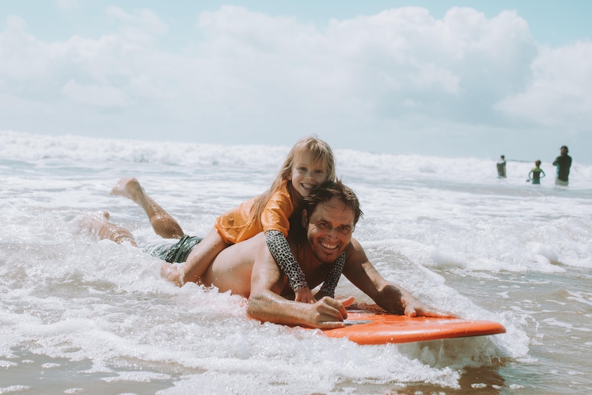 A smiling young girl in an orange rashie on the back of a man who is riding an orange surfboard on his stomach.