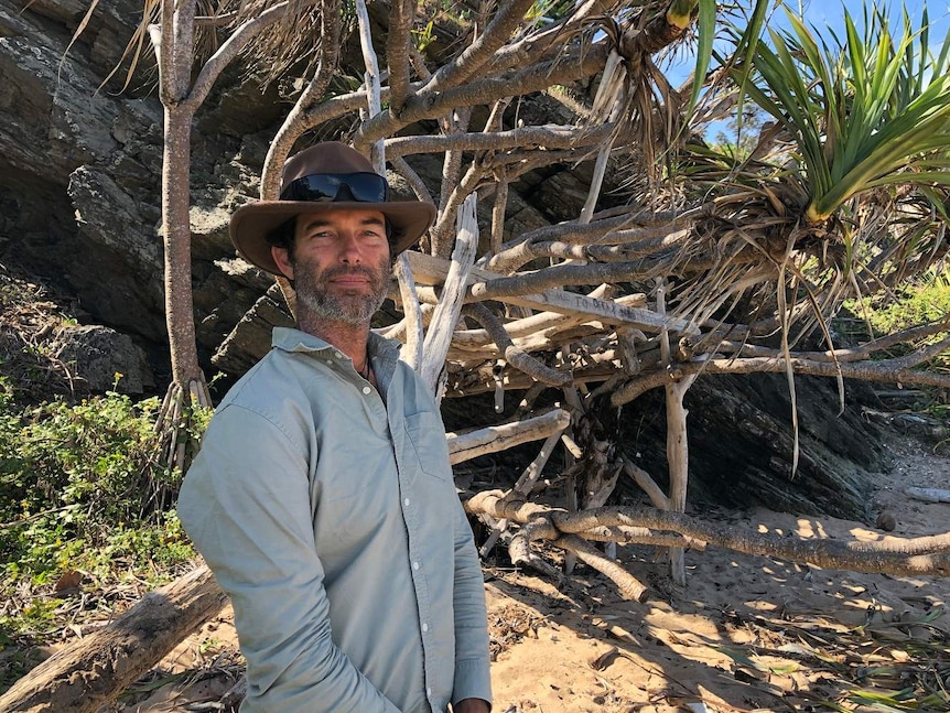 A man in a shady hat and dressed in outdoor working gear stands in front of a patch of pandanus trees