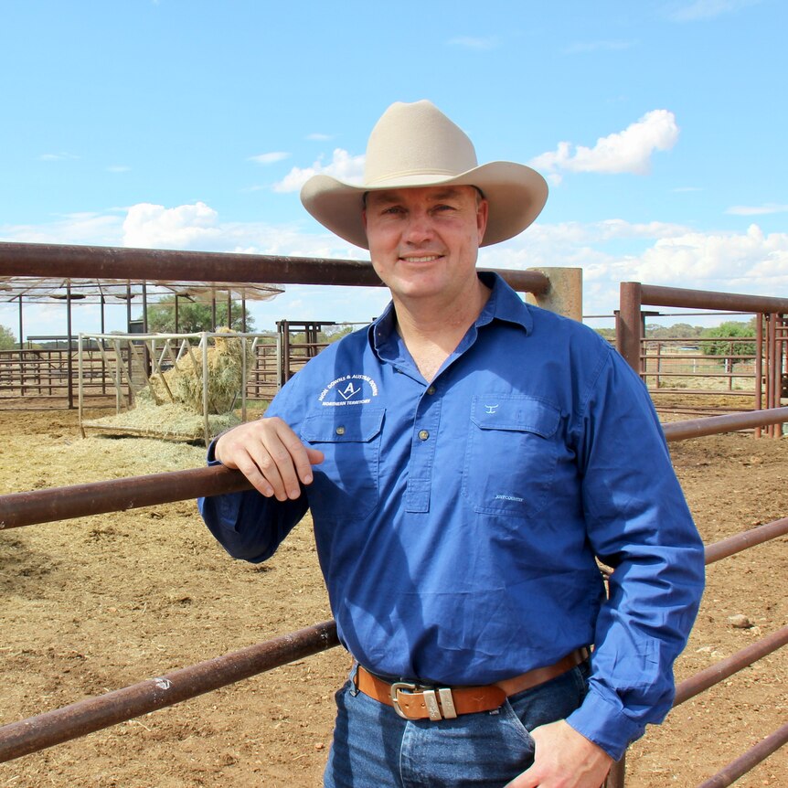 a man in a blue shirt and white broad-brimmed hat leaning on a cattle yard rail.