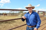 a man in a blue shirt and white broad-brimmed hat leaning on a cattle yard rail.