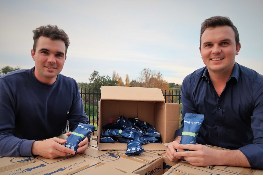 Two men pose with a cardboard box of dog kibble and smile down the camera