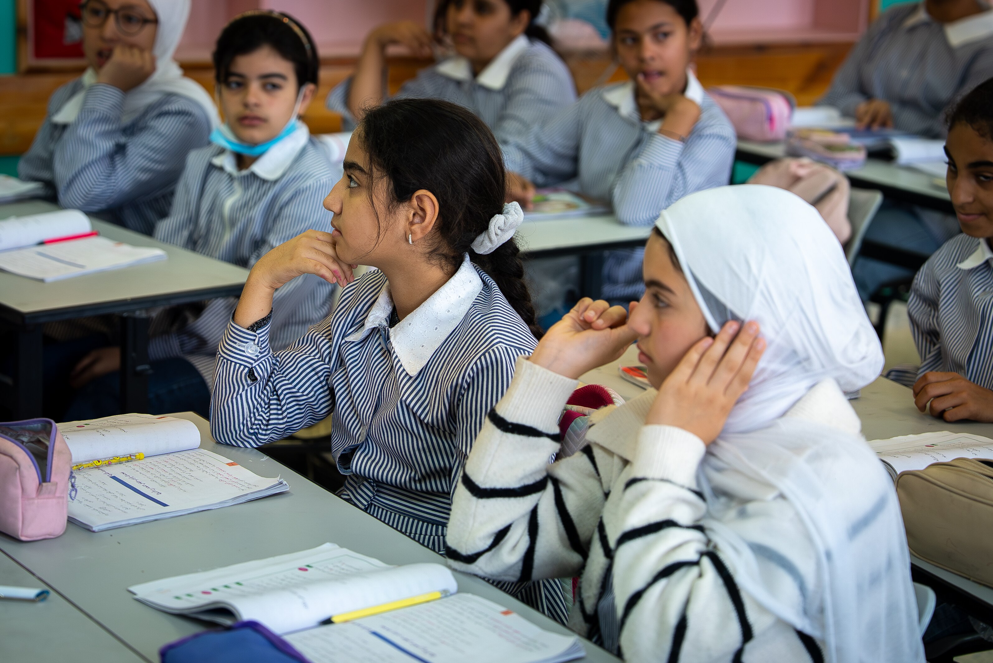 Two girls wearing hijabs sit in a classroom with their books open on their desks.