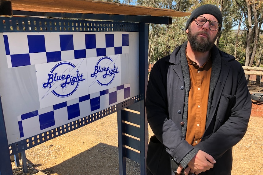 a man wearing a beanie and overcoat stands by a sign of Blue Light Youth camp.