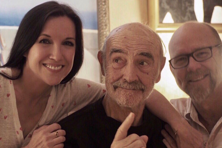 An elderly man grins while a woman and a man put their arms around his shoulders.