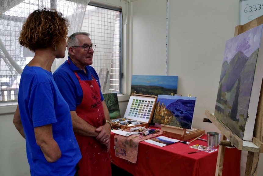 A woman, with one arm amputated, stands talking to a man, looking at an art work.