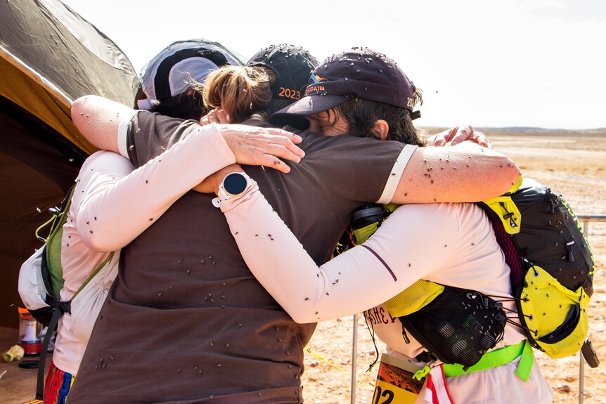 Three women embrace after finishing a run, covered in flies.