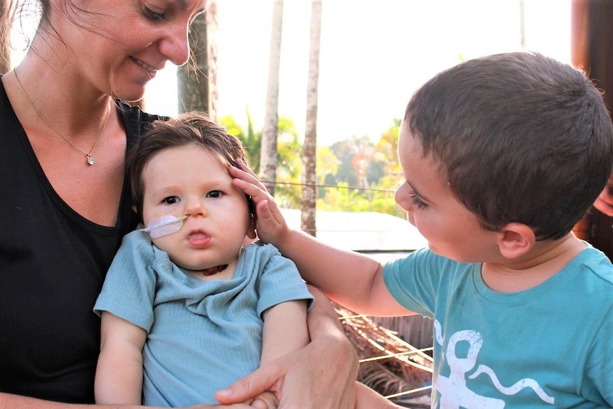 Young boy wearing blue shirt gently touches head of baby brother on verandah at sunset in Far North Queensland.