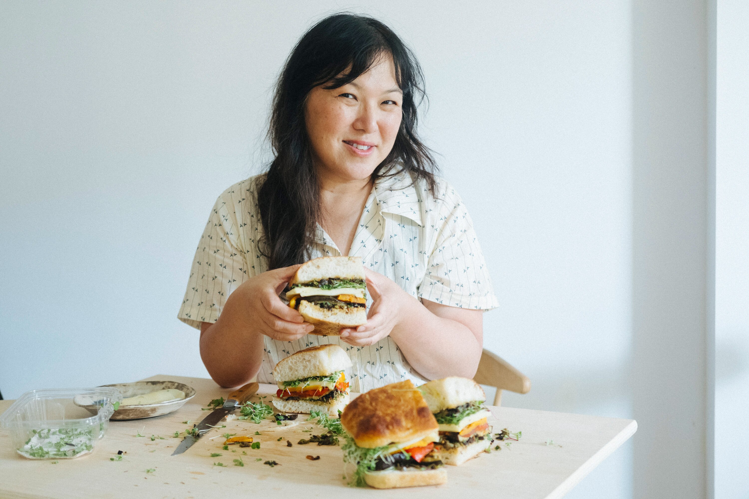 Food writer and cookbook author Hetty Lui McKinnon holding a roasted veg and cheese sandwich at a timber table.