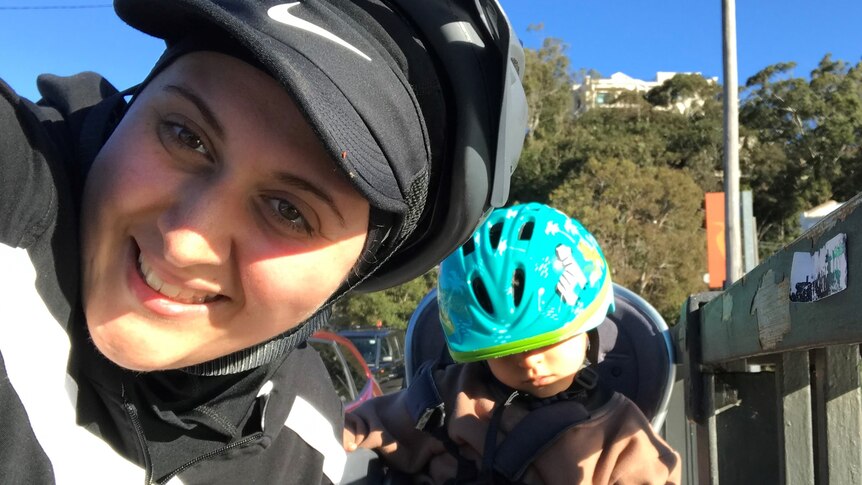 Selfie of a woman smiling in a baseball cap and helmet, with a child in a helmet. 