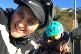 Selfie of a woman smiling in a baseball cap and helmet, with a child in a helmet. 