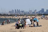 People gather on a bayside Melbourne beach under sunshine, with the Melbourne CBD skyline visible in the background.