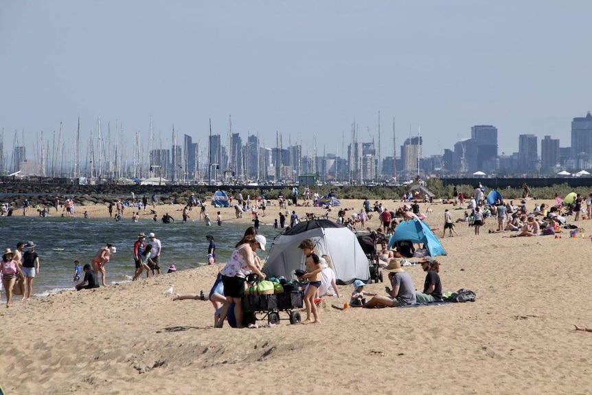 People gather on a bayside Melbourne beach under sunshine, with the Melbourne CBD skyline visible in the background.