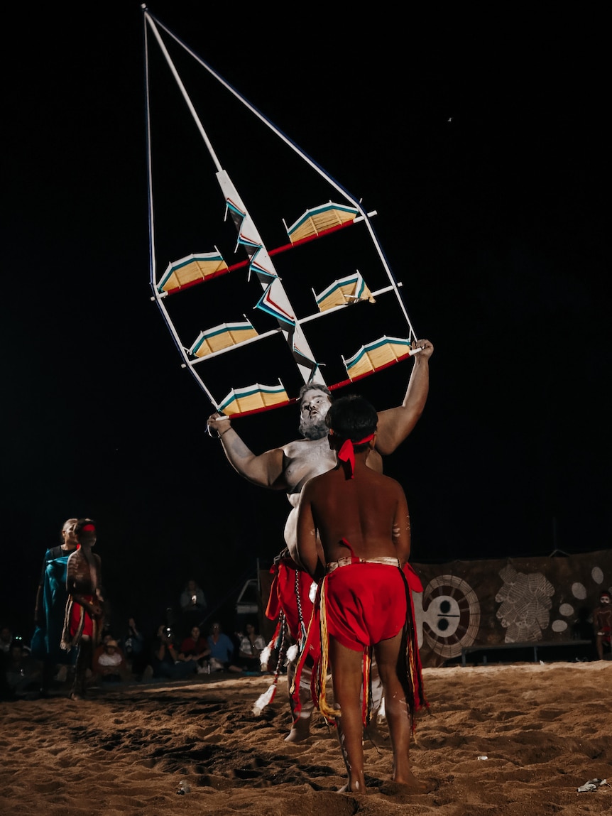 A man performs a traditional Aboriginal dance, holding a large colourful totem above his head.